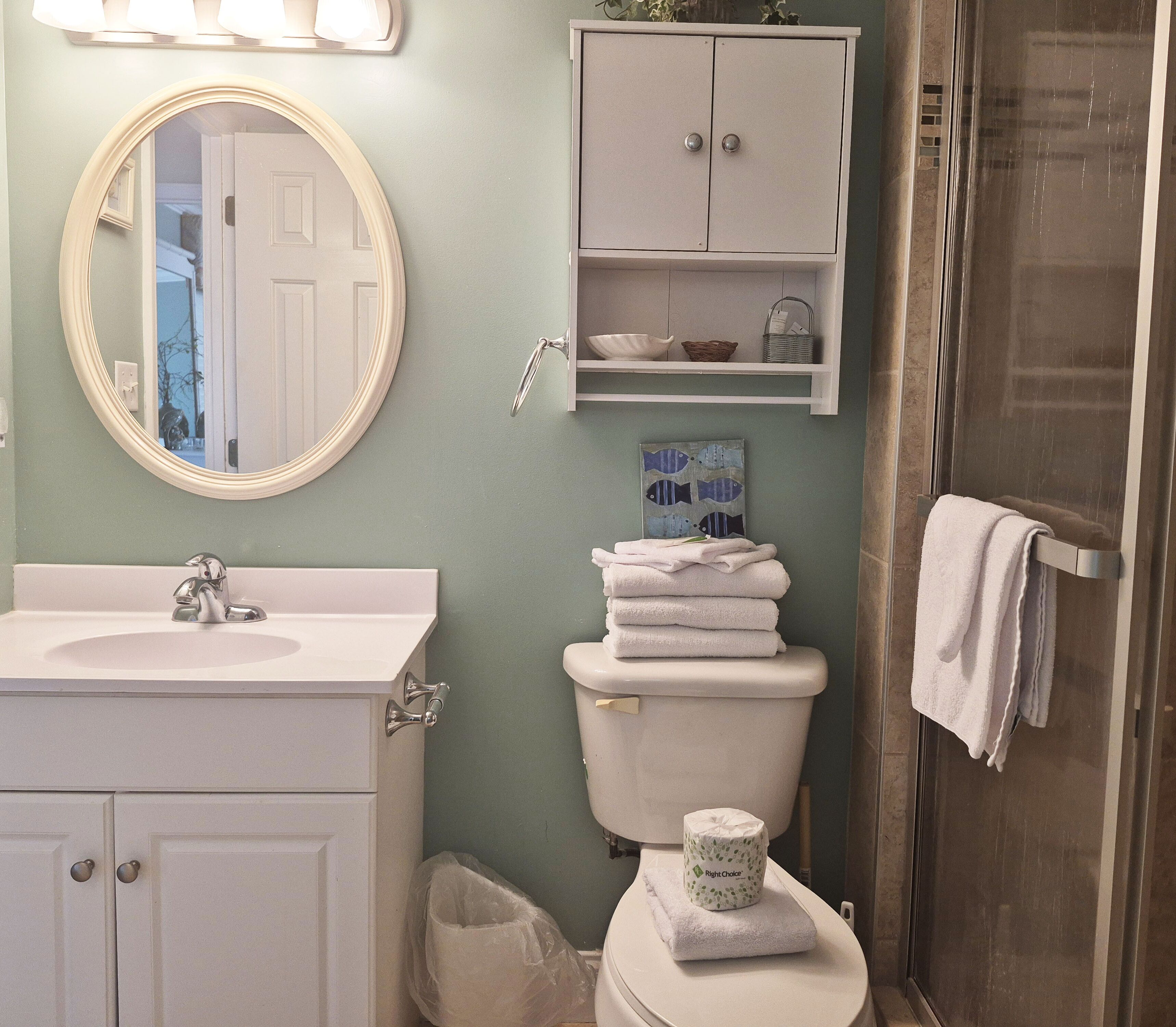 A bathroom with a sink, mirror, toilet, and shower. There are towels stacked on the toilet and a trash can.