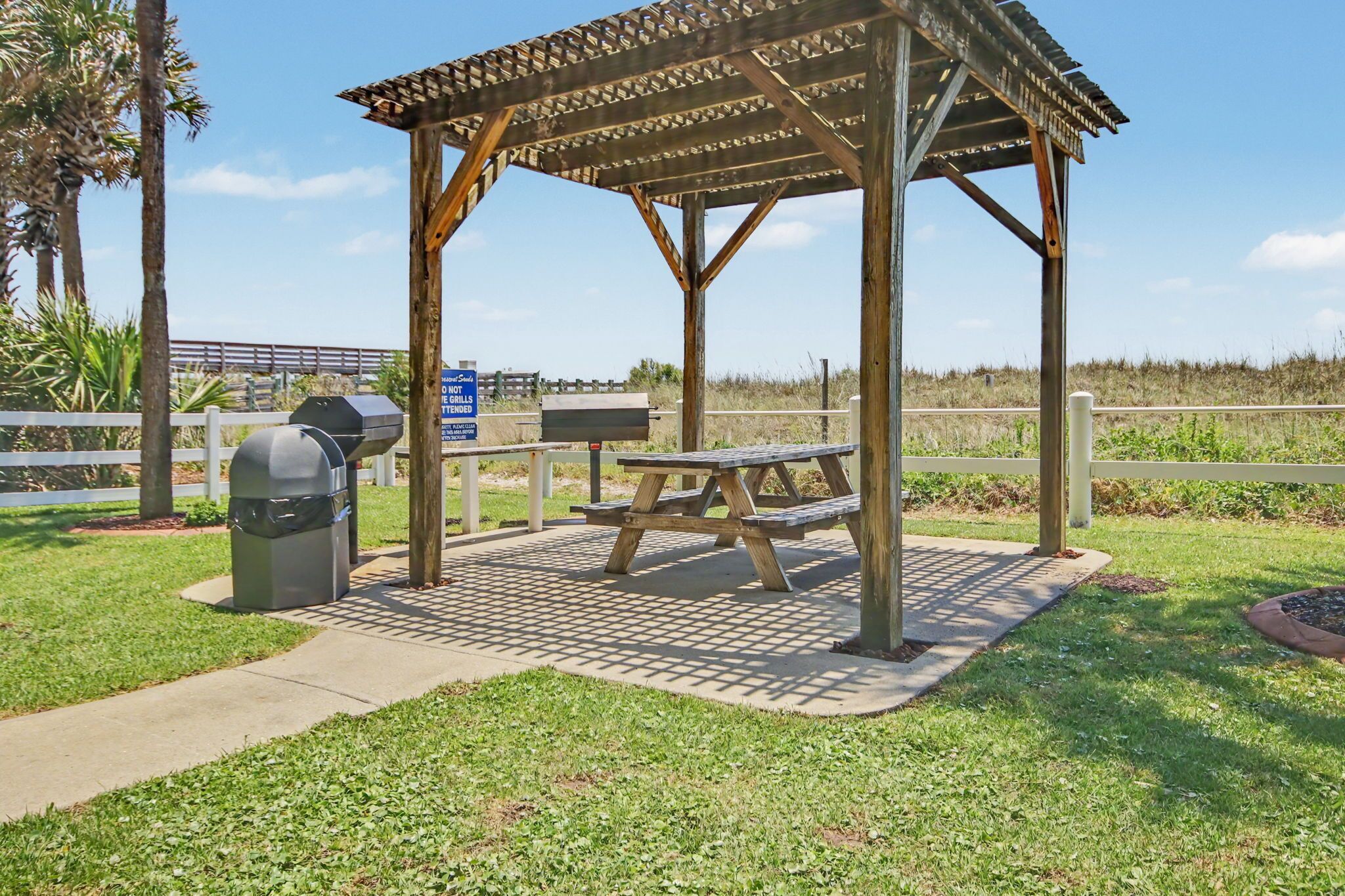 A gazebo with a picnic table and grill, surrounded by grass and a fence.