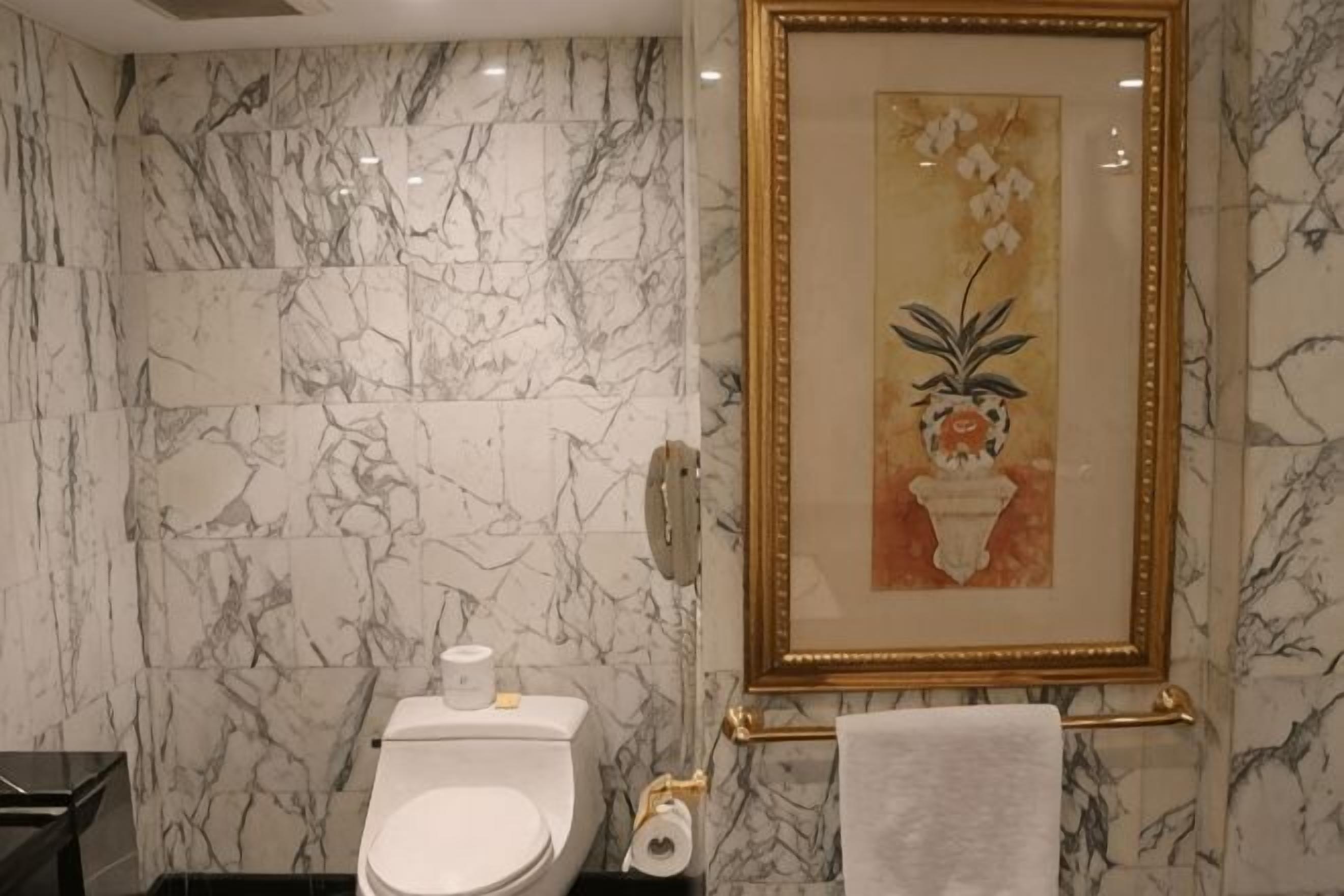 Un baño con paredes de mármol, un inodoro, un cuadro enmarco de flores y un perchero para toallas.