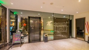 Un vestíbulo de hotel con diseño moderno, que presenta un mostrador de recepción, un separador de vidrio con un mural decorativo y una pequeña disposición de plantas.