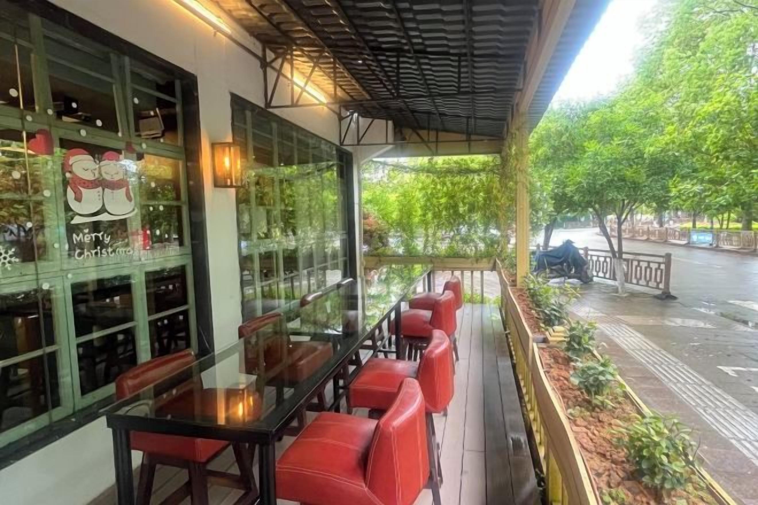 Un espacio al aire libre con mesas y sillas, un cerramiento de vidrio con botellas y vista a una calle con árboles.