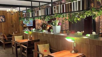 Interior de restaurante con mesas y sillas de madera, una barra con lámpara verde y una pared decorada con plantas y libros.
