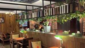 Un interior de restaurante con mesas y sillas de madera, una barra con una planta en maceta y una pared con estanterías y vegetación.