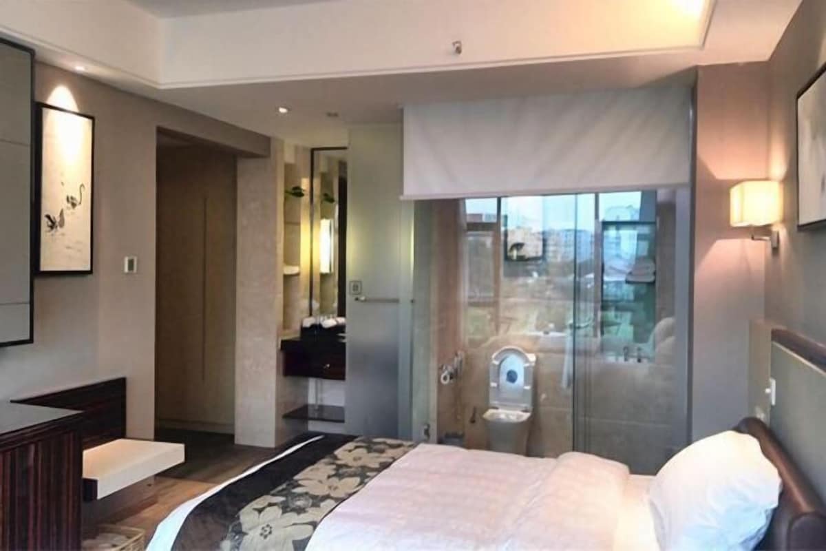 A hotel room with a bed, a bathroom with a glass shower, a vanity, and a view of the city.