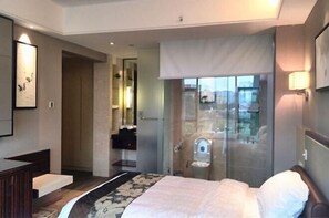 A hotel room with a bed, a bathroom with a glass shower, a vanity, and a view of the city.