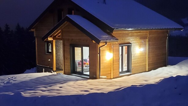 A wooden cabin with a snow-covered roof, a front porch, and a lit interior visible through the windows. - Chalet "DIANA", at the foot of the ski slopes. Pets allowed  (Gérardmer)