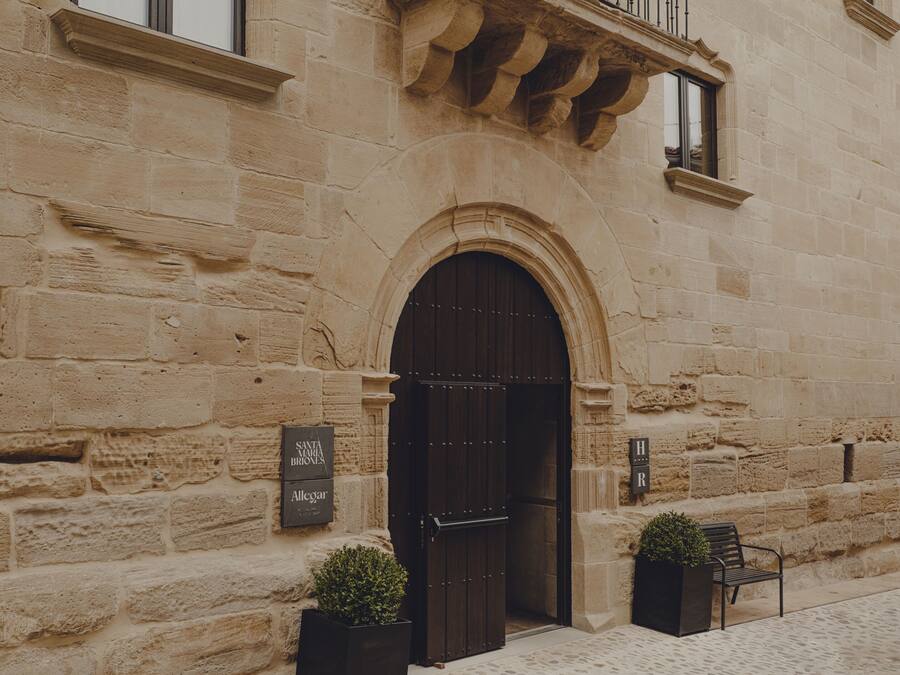 Un edificio de piedra con un amplio acceso en arco, un balcón con barandilla de hierro forjado y dos macetas con plantas en la entrada.