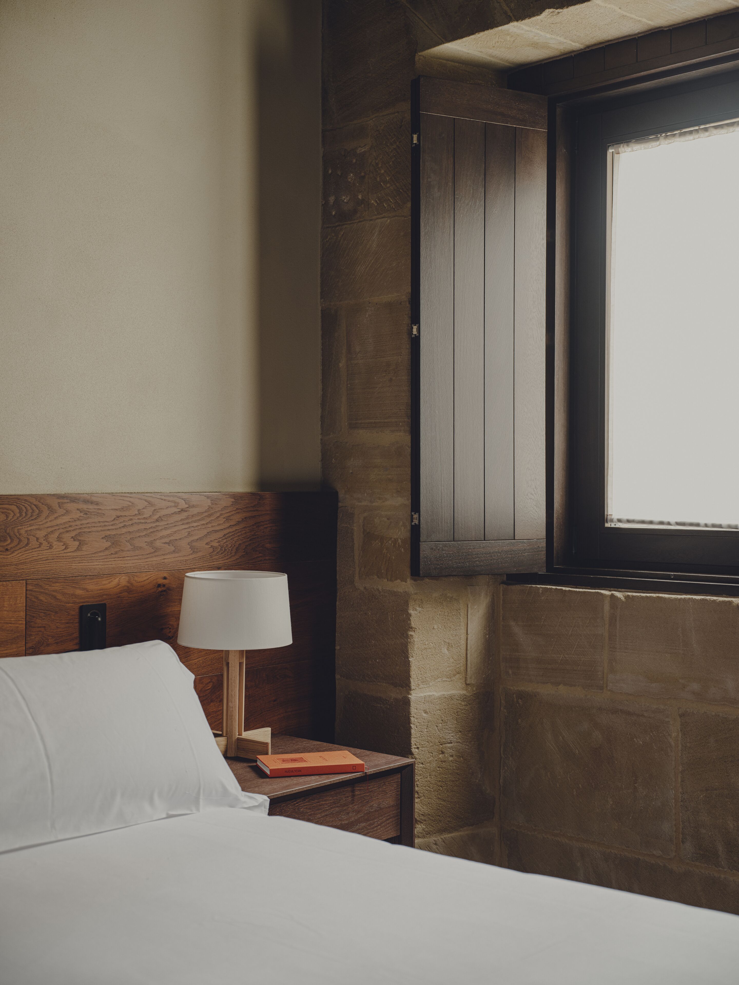 Habitación de hotel con cabecera de madera, lámpara de noche, mesita de noche pequeña y ventana con vistas.