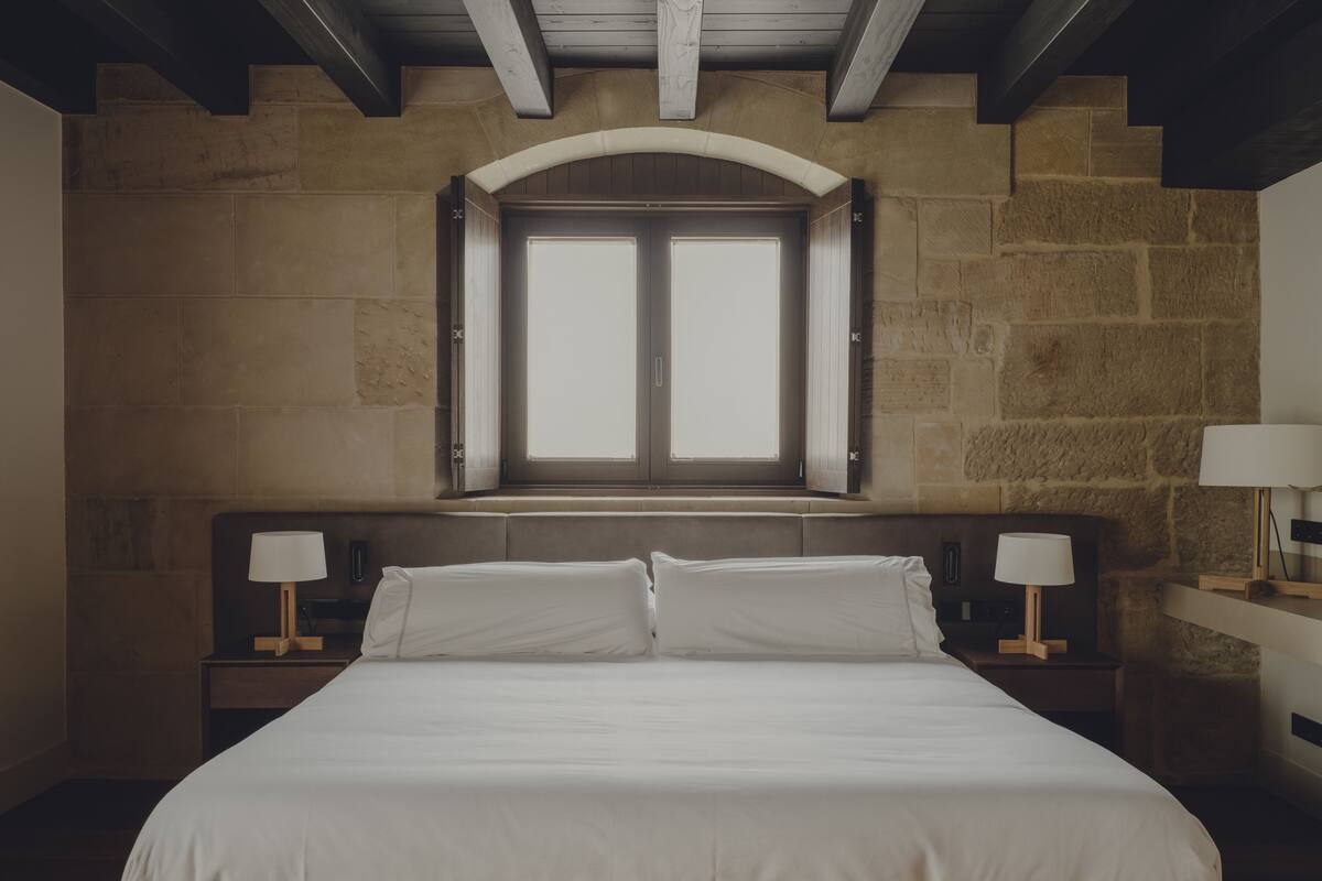 A bedroom with a large bed, two bedside tables with lamps, a stone wall, and a window above the bed.