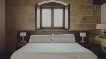A bedroom with a large bed, two bedside tables with lamps, a stone wall, and a window above the bed.