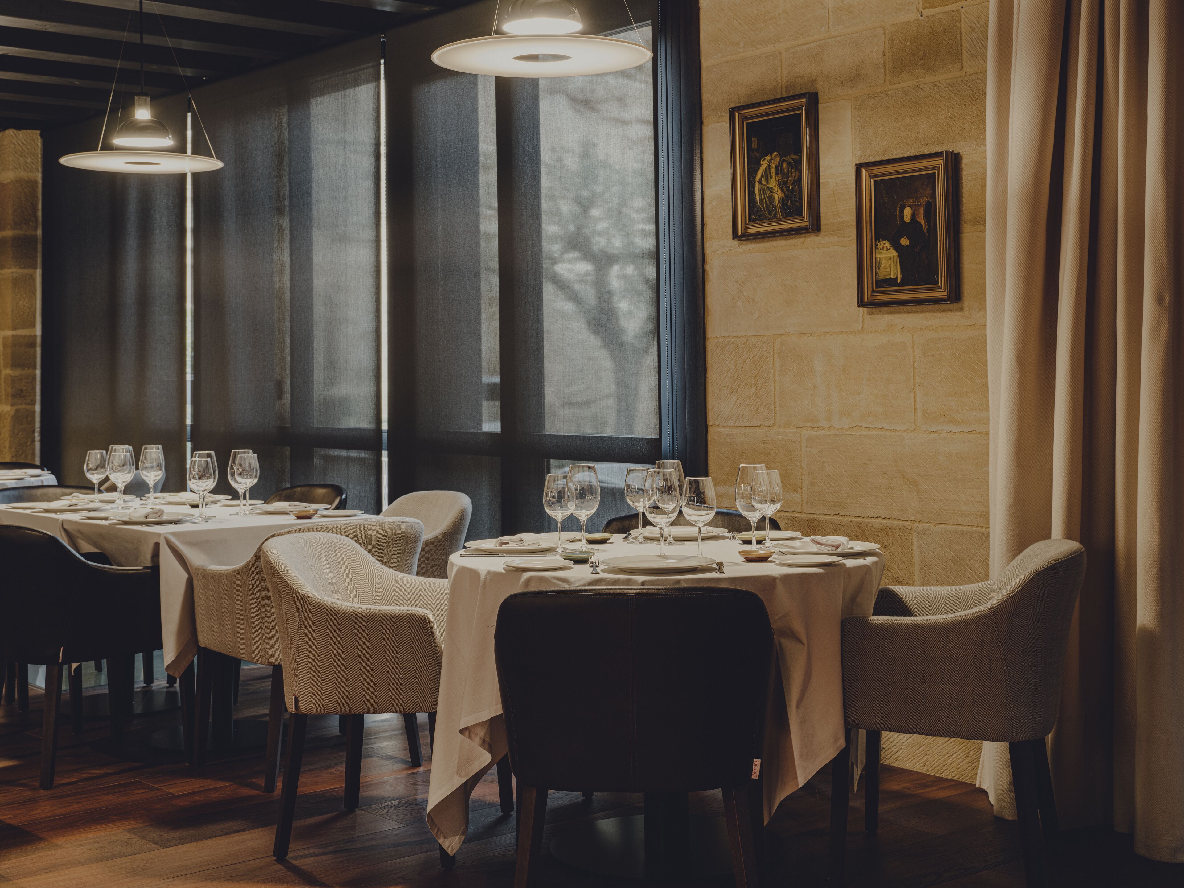 Een restaurant met meerdere tafels voor diners, grote ramen en hangende pendantlampen.