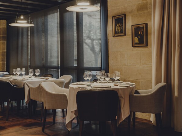 Un restaurante con varias mesas dispuestas para comer, amplias ventanas y lámparas colgantes.