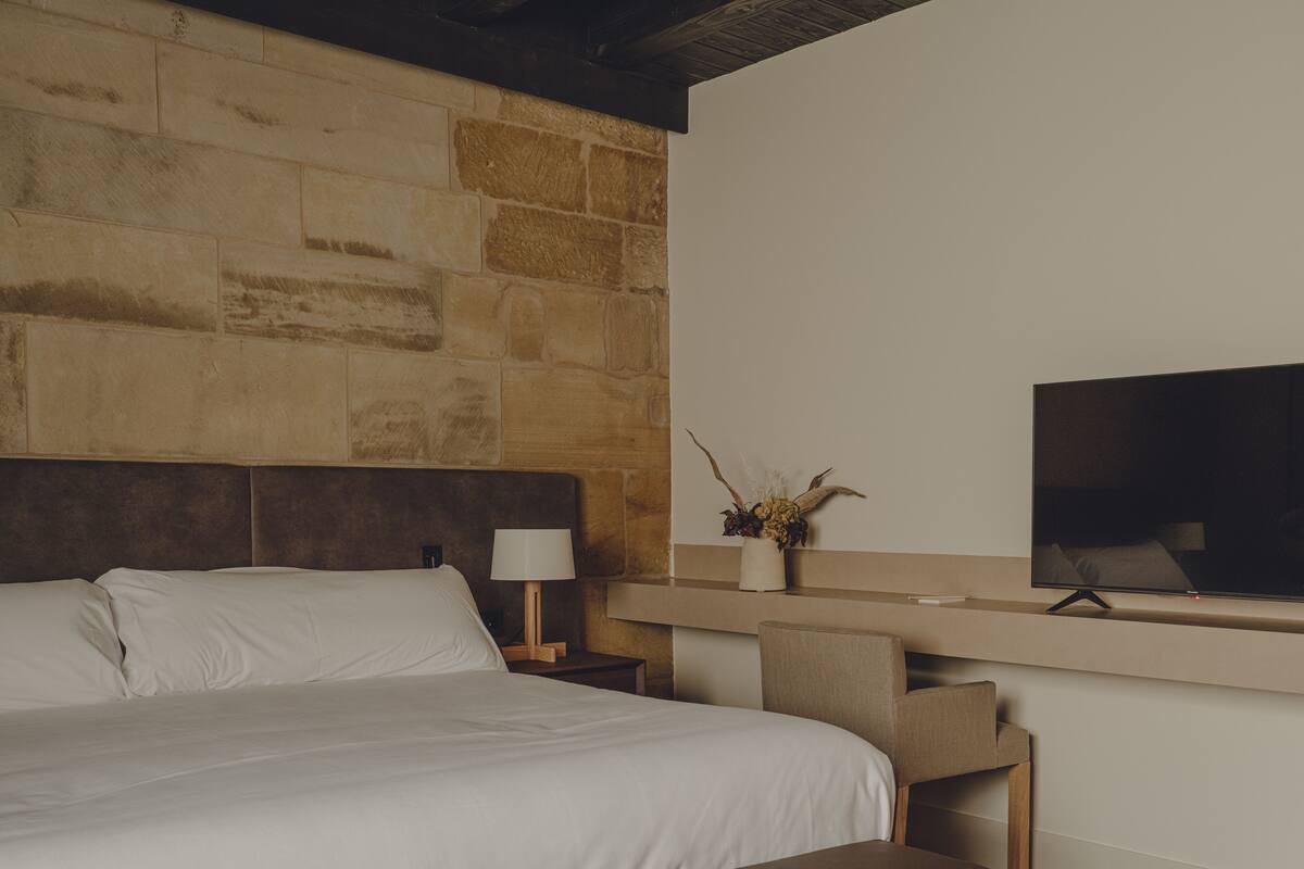 A modern hotel room with a stone accent wall, a large bed with white linens, a bedside table with a lamp, a flat-screen TV mounted on the wall, and a wooden bench.