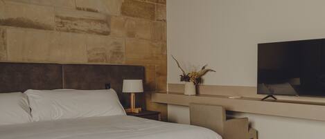 A modern hotel room with a stone accent wall, a large bed with white linens, a bedside table with a lamp, a flat-screen TV mounted on the wall, and a wooden bench.