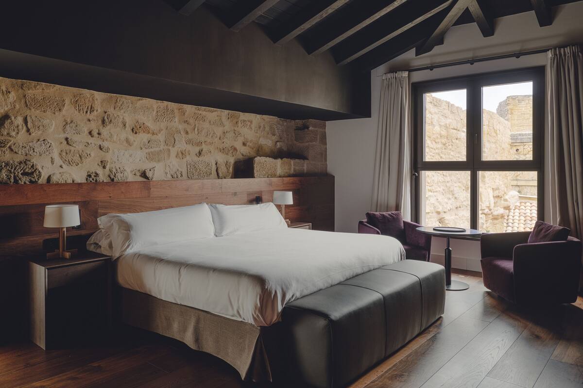 A bedroom with a stone wall, wooden ceiling beams, a large bed, two bedside tables, a chair, and a window with curtains.