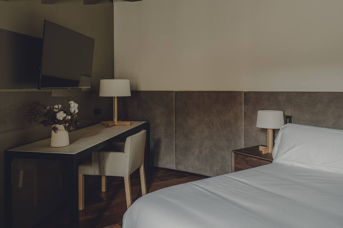 A hotel room with a bed, a desk with a chair, a TV mounted on the wall, and a nightstand with a lamp.
