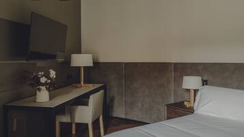 Quarto de hotel com uma cama, uma mesa com cadeira, uma televisão na parede e uma mesinha de cabeceira com abajur.