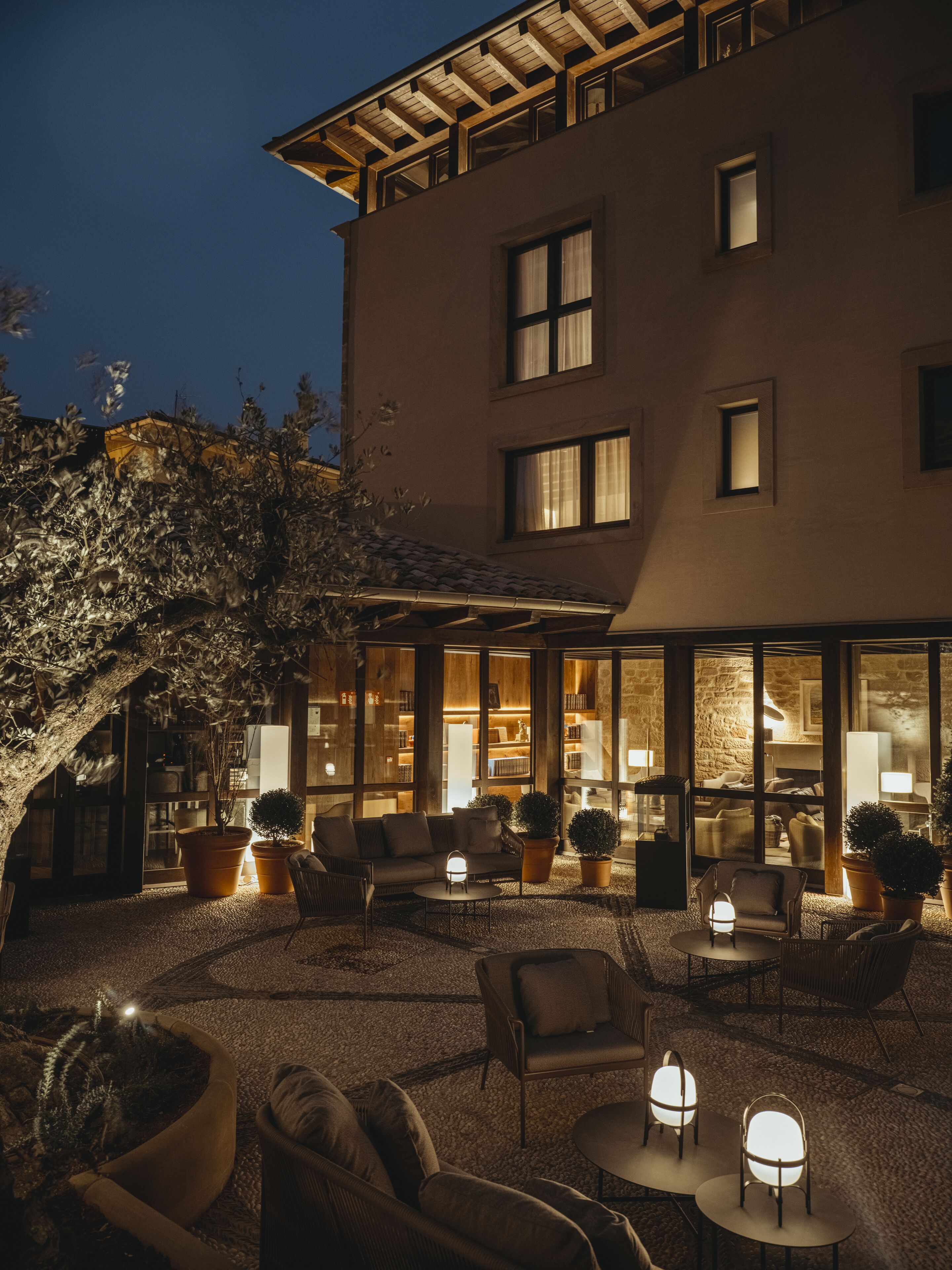 A building with a covered outdoor seating area, furnished with sofas and tables, and surrounded by potted plants.