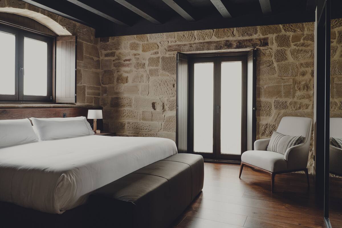 A bedroom with a large bed, a chair, a nightstand, and a stone wall.