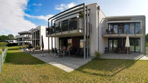 A modern two-story house with a patio area, outdoor seating, and a large window overlooking the garden.