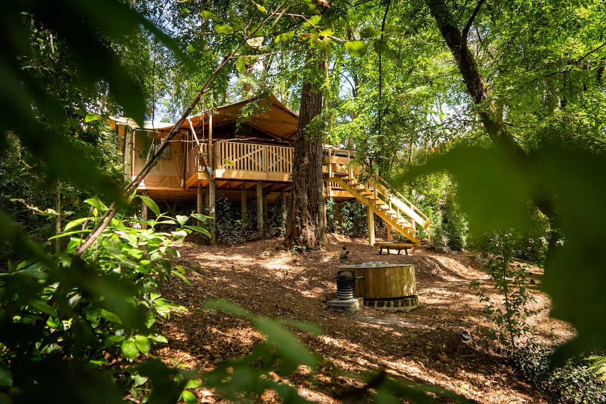 A wooden cabin with a deck and staircase, surrounded by trees and a hot tub.