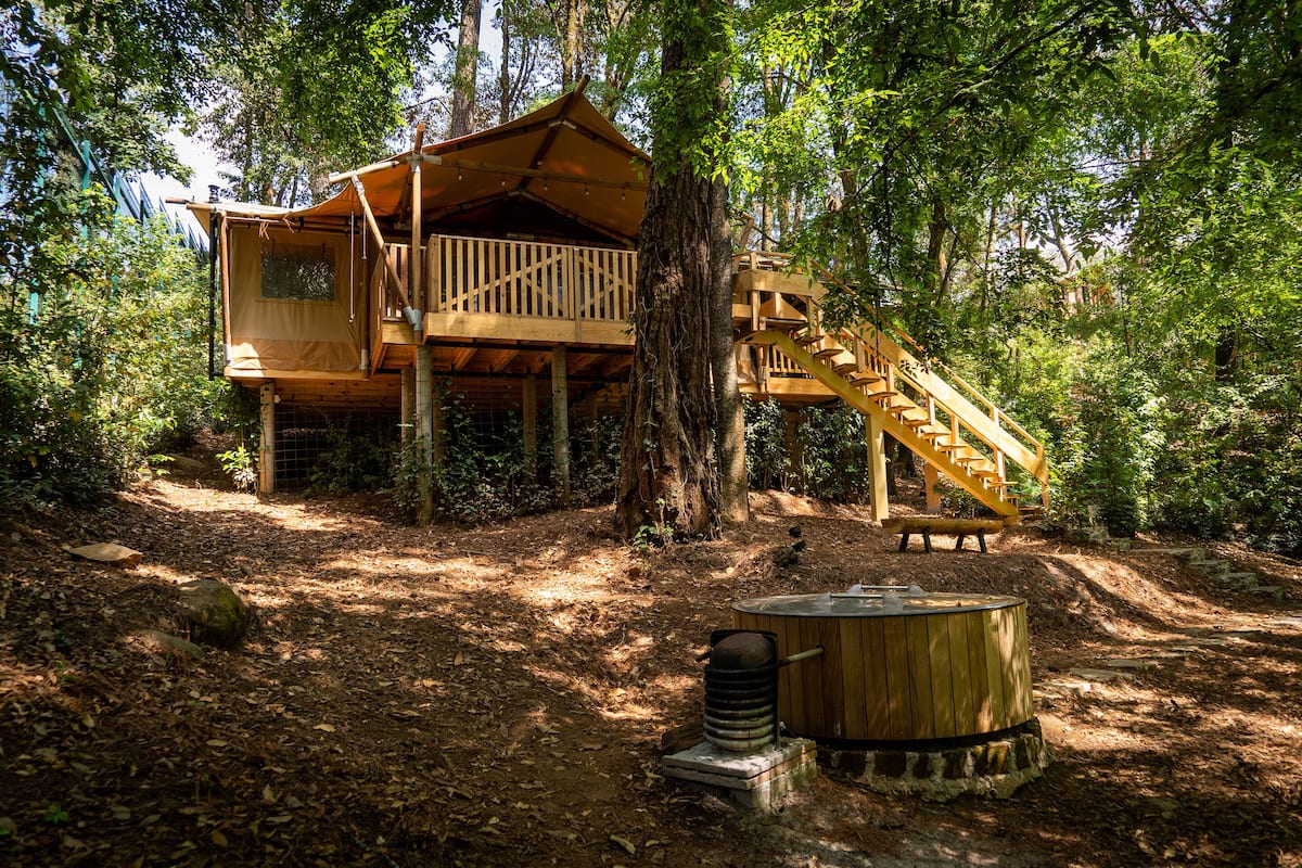 A wooden treehouse with a balcony and a staircase, surrounded by trees and a hot tub.