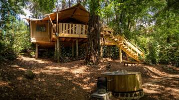 A wooden treehouse with a balcony and a staircase, surrounded by trees and a hot tub.