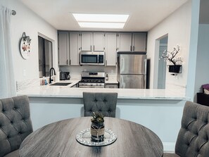 Een moderne keuken met grijze kastjes, een roestvrijstalen koelkast, een magnetron en een fornuis. Er staat een eettafel met een bloevas en een decoratief bord.