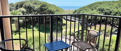Un balcón con dos sillas de mimbre y una mesita, con vistas a un jardín exuberante y una vista distante del mar.