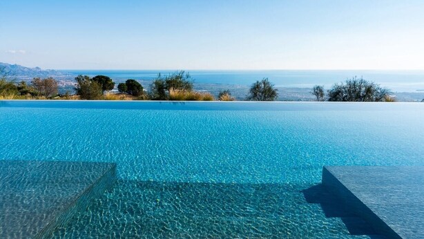 Una piscina a sfioro di colore blu intenso, con vista sul mare e montagne in lontananza.
