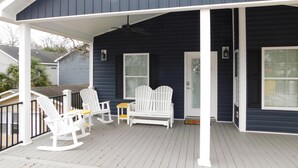 A covered porch with white rocking chairs and a small table, a white door with a glass panel, and a dark blue building. - V29 Family-friendly 3BR w/ Outdoor Kitchen + 2 Lounges, Limo Golf Cart (Myrtle Beach)