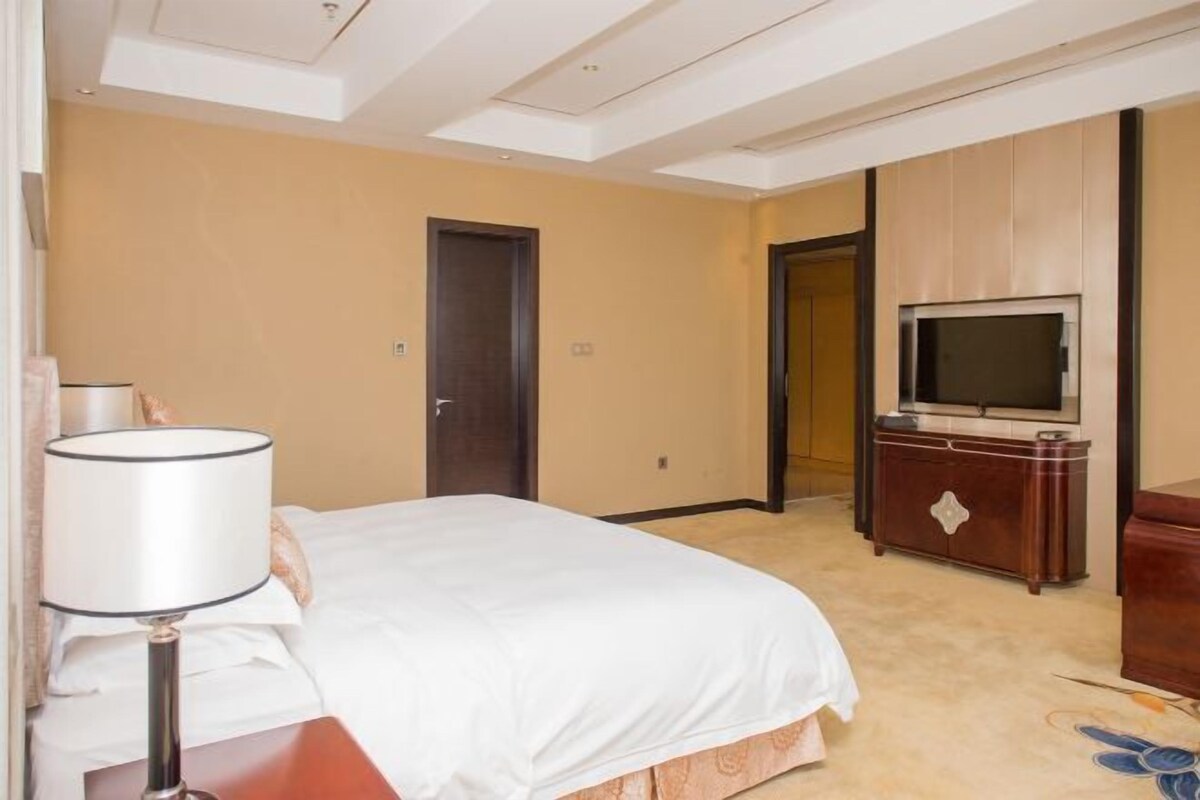 Habitación de hotel con cama, televisión sobre un mueble de madera, lámpara y puerta que da a otra habitación.