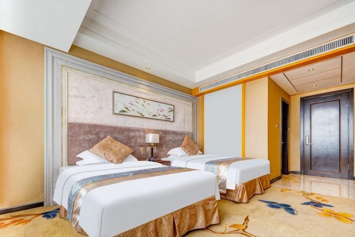 A hotel room with two beds, a large painting on the wall, a carpet with a floral pattern, and a door.