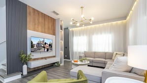 Ein modernes Wohnzimmer mit einem an einer Holzvertäfelung montierten Flachbildfernseher, einem grauen Ecksofa, einem grünen Sessel und einem weißen Couchtisch mit Dekoration.