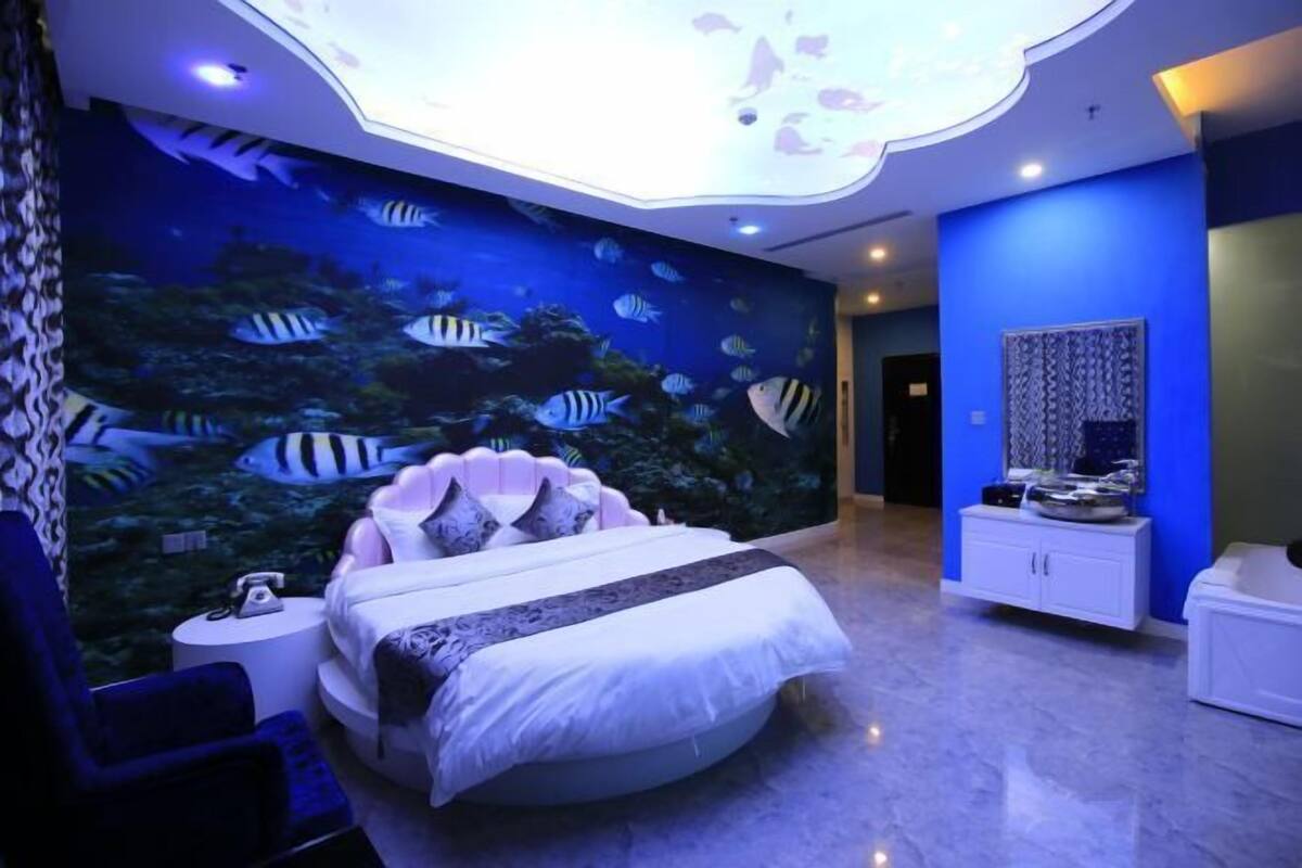A bedroom with a large aquarium wall, a round bed, a nightstand, and a bathroom with a mirror and sink.