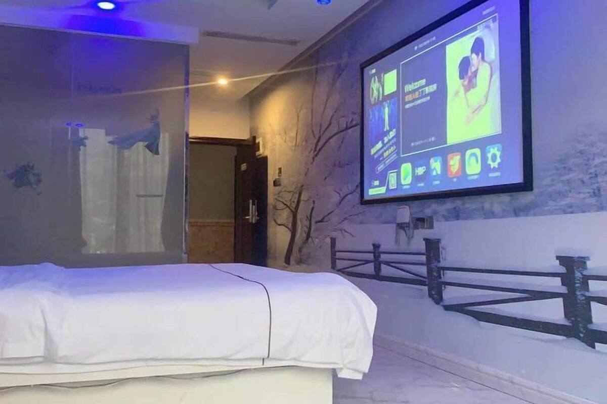 A hotel room with a bed, a wall-mounted TV displaying a welcome screen, a glass partition, and a view of a snowy outdoor area.