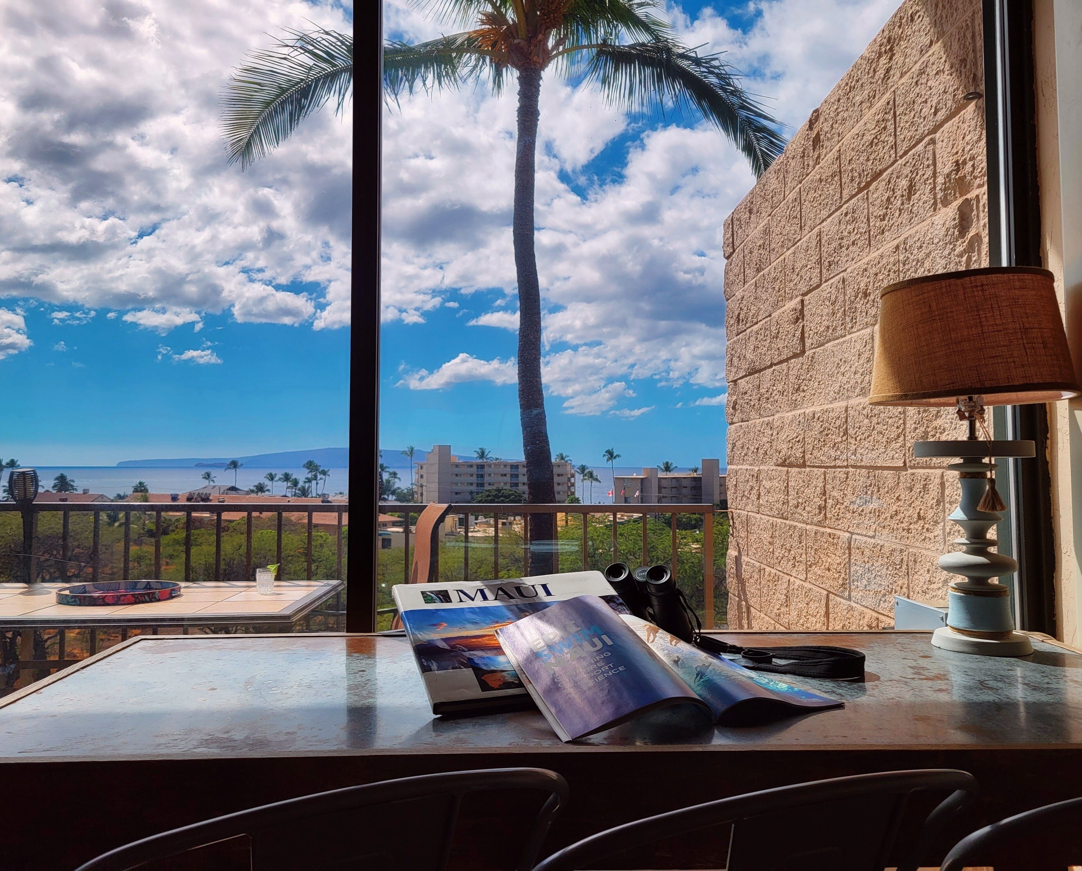 Un balcón con vista al océano y palmeras, una mesa con una revista y una lámpara.