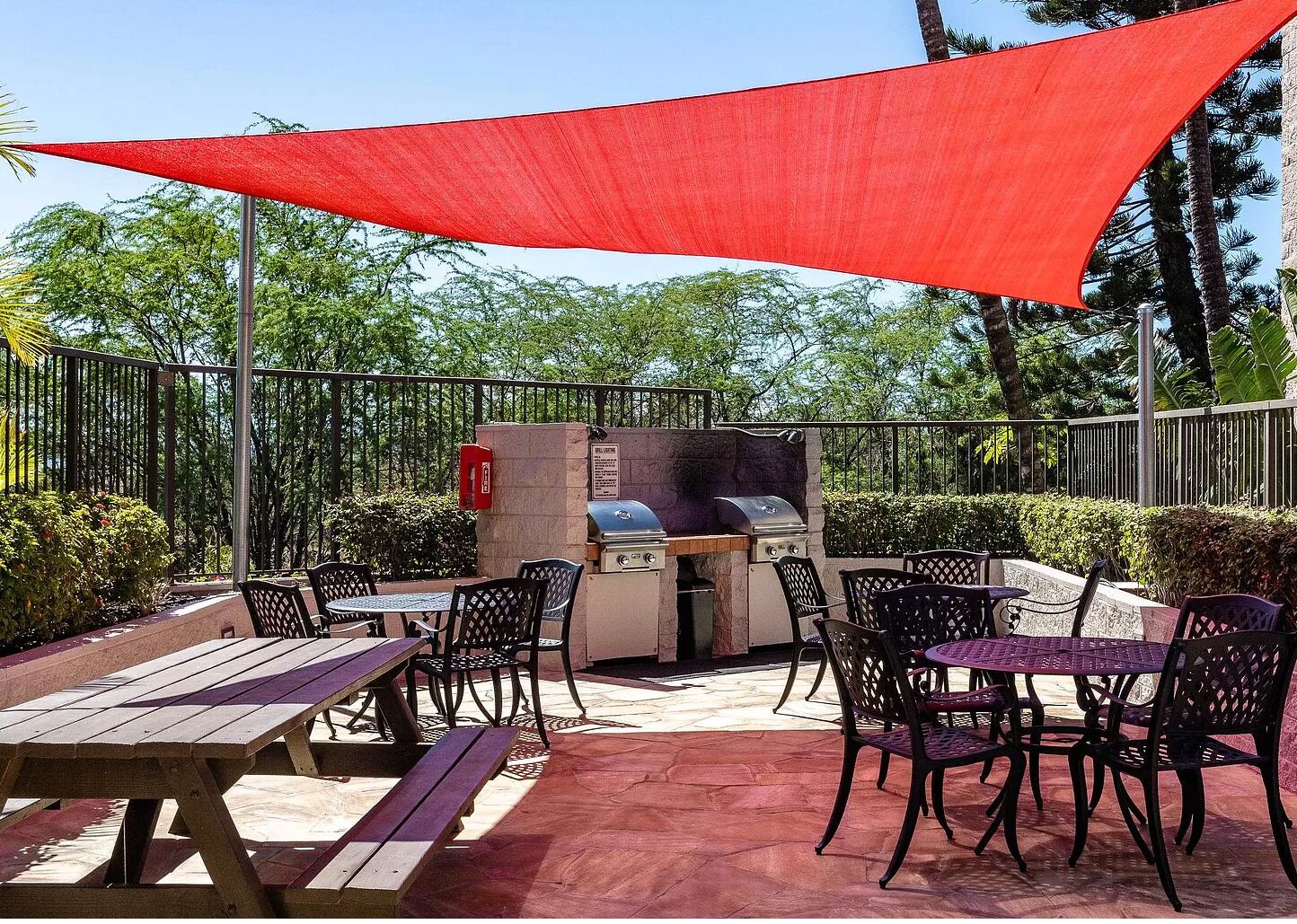 An outdoor patio with picnic tables and chairs, a barbecue grill, and a red shade sail.