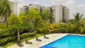 Una piscina con sillones de descanso, rodeada de palmeras y un edificio de varios pisos al fondo.
