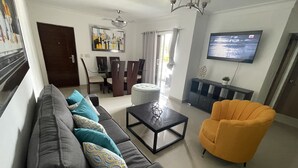 Una sala de estar con sofá, una mesa de centro, un sillón amarillo, televisor, comedor y candelabro.