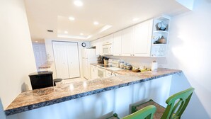 A kitchen with white cabinets, a granite countertop, and a microwave. There are two green chairs and a small shelf with decorative items. - Oceanfront 2BR 2BA Condo 5th Floor Balcony Ocean Views, 504 (Indian Harbour Beach)