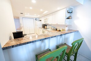 A kitchen with white cabinets, a granite countertop, and a microwave. There are two green chairs and a small shelf with decorative items.