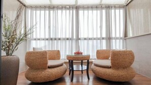 Een gezellig zithoekje met twee geweven stoelen en een klein rond tafeltje, een groot raam met dunne gordijnen en een potplant aan de linkerkant.