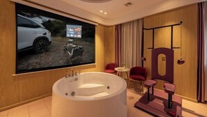 Een moderne badkamer met een groot bad, een televisiescherm waarop een auto wordt getoond, en een klein zitgedeelte met twee stoelen.