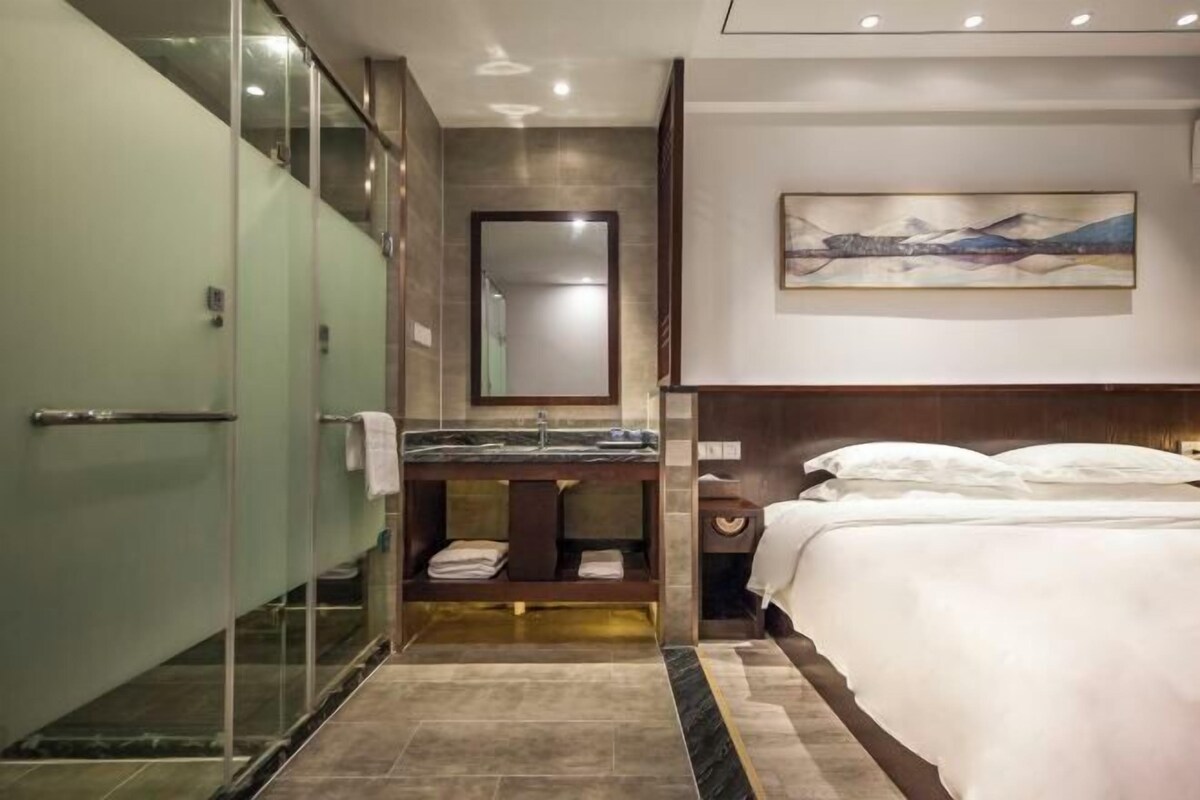 Ein modernes Hotelzimmer mit Duschkabine aus Glas, Marmorboden, einem großen Bett mit weißer Bettwäsche, einem Nachttisch aus Holz und einem Gemälde an der Wand.