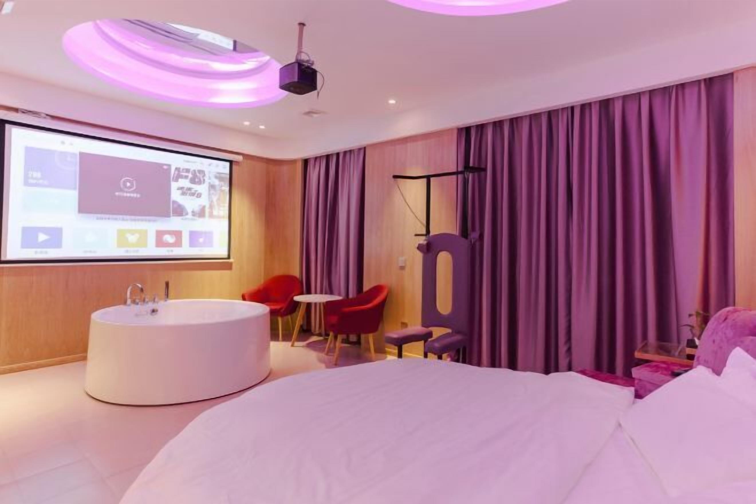 A modern hotel room with a large bed, a round bathtub, a small table, and a projector screen displaying a video.