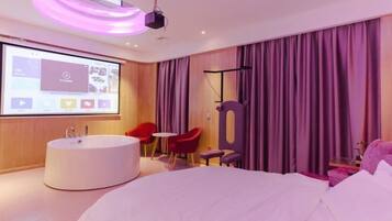 A modern hotel room with a large bed, a round bathtub, a small table, and a projector screen displaying a video.