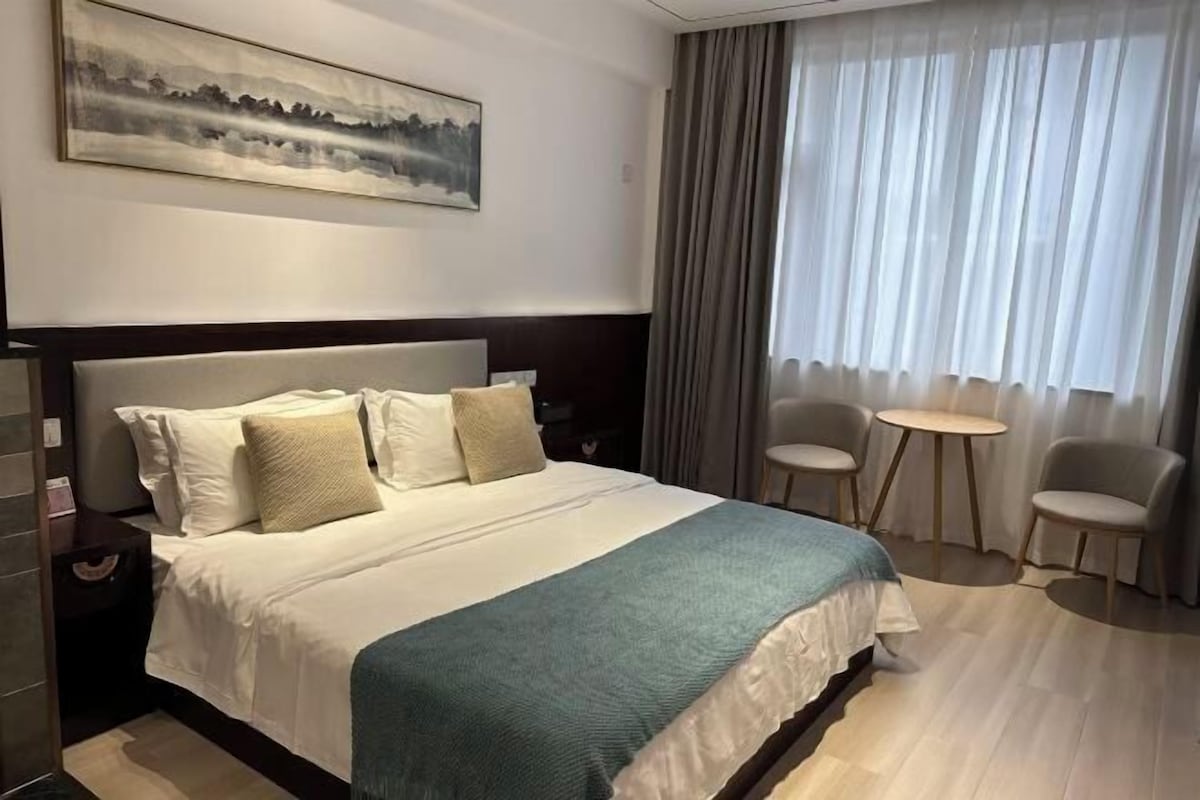 Ein Hotelzimmer mit einem großen Bett, einem Bild an der Wand, einem Fenster mit Vorhängen und zwei Stühlen am Fenster.