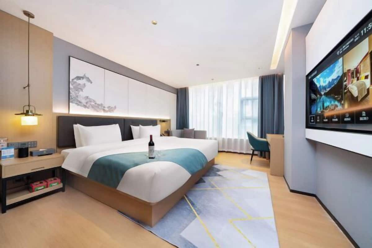Una habitación de hotel moderna con una cama grande, un escritorio, una silla, un televisor y un amplio ventanal con cortinas.