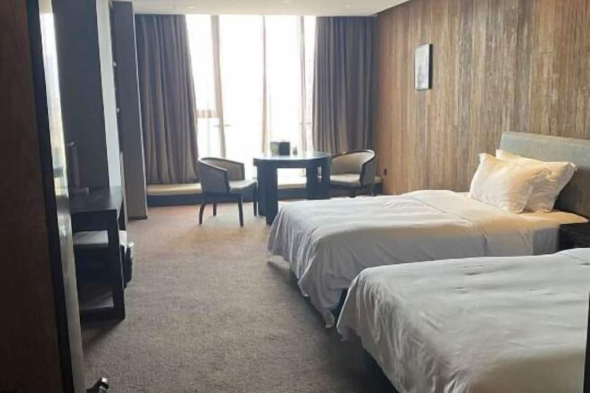 A hotel room with two beds, a small table, a chair, and a view of the outdoors.